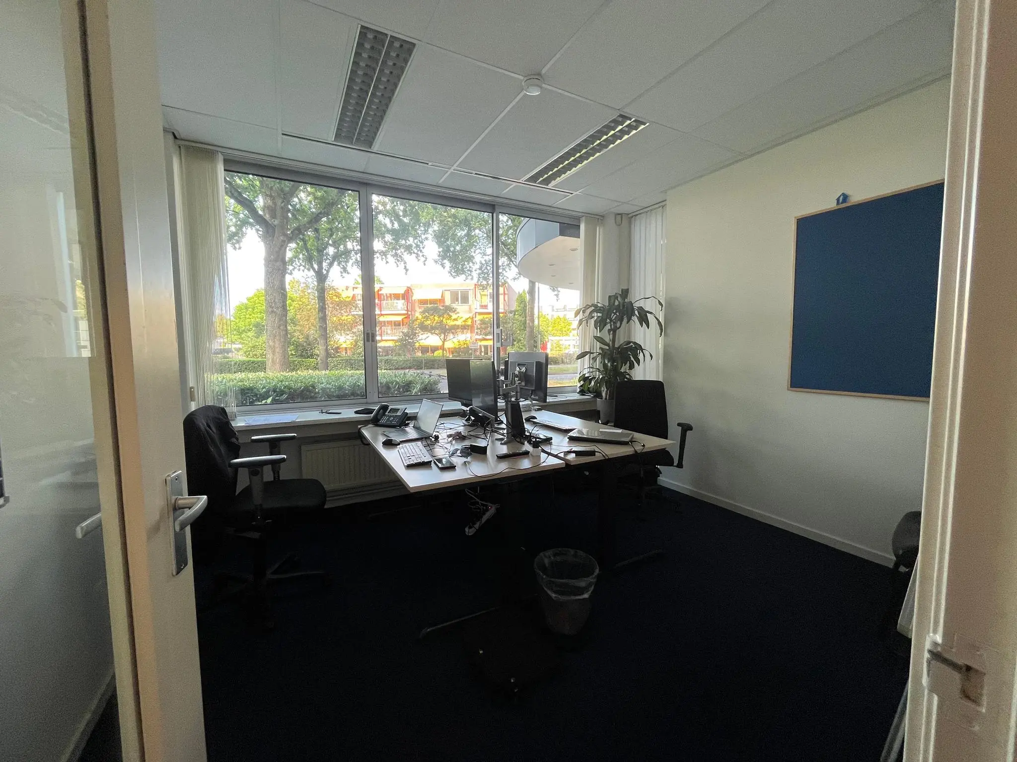 Kleine kantoorruimte aan de Jan van Eycklaan met bureaus, stoelen, computers en een groot raam met uitzicht op bomen en een woongebouw.